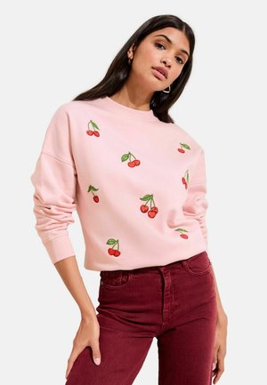 Woman wearing a light pink sweatshirt with cherry embroidery and high-waisted dark red pants against a plain background.