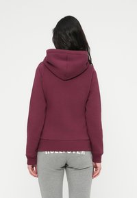 Μπορντό φούτερ με κουκούλα, εφαρμοστή σχεδίαση και ριμπ μανσέτες. Φορεμένο με γκρι κολάν που διαθέτουν λεπτομέρεια λογότυπου στη μέση.