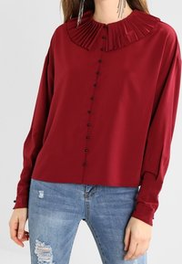 Blusa bordeaux con colletto plissettato, chiusura con bottoni frontali e maniche lunghe. Abbinata a jeans chiari strappati.