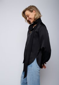 Czarna satynowa koszula o luźnym kroju, z wysokim kołnierzem i detalami w postaci guzików, zestawiona z jasnoniebieskimi dżinsami. Gładka tekstura, płynny fason.