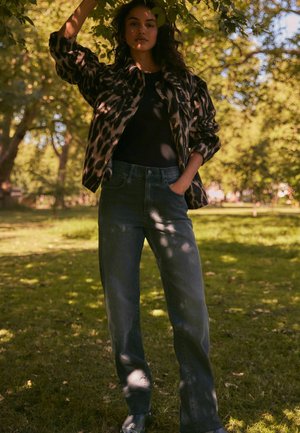 Jasje met luipaardprint, zwart shirt, hoge taille donkere denim jeans, relaxed fit, staand tussen groen, zonlicht dat door de bomen heen komt.
