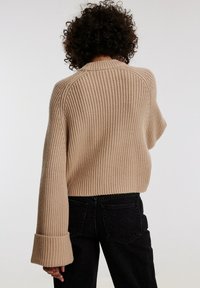 Suéter de punto beige con diseño acanalado, corte cropped, puños anchos y ajuste relajado, combinado con pantalones negros de tiro alto.