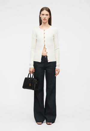 Vrouw draagt een wit, met knopen gesloten, top met lange mouwen, zwarte wijde jeans en houdt een zwarte handtas vast, staand tegen een effen achtergrond.