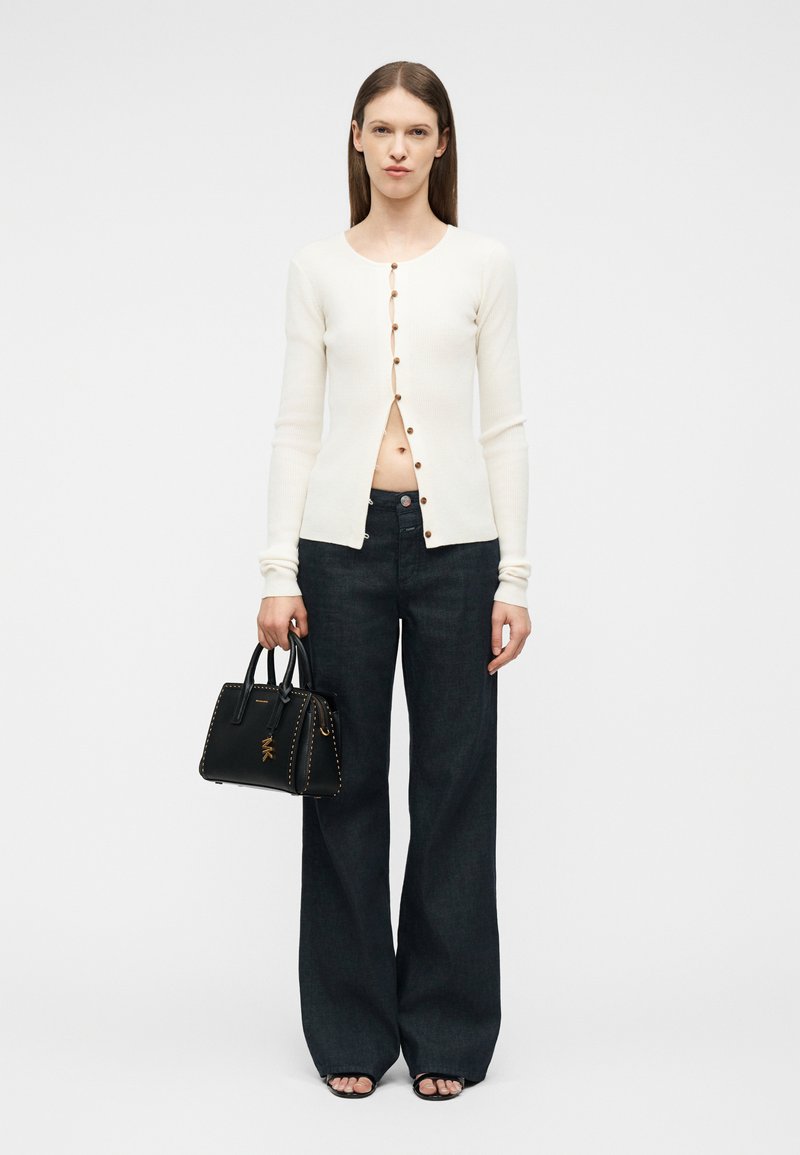 Γυναίκα με λευκό μακρυμάνικο πουκάμισο με κουμπιά, μαύρο τζιν παντελόνι με φαρδιά μπάρα, κρατώντας μια μαύρη τσάντα, στέκεται μπροστά σε απλό φόντο.