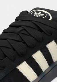 Melns sporta apavu pāris ar teksturētām gumijas šņorēm, zamša virsmu, krēmkrāsas svītrām un apaļu logotipu uz mēlītes, ar gludu pirksta daļu.