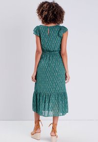 Robe verte à motifs floraux avec de courtes manches volantées, une taille ceinturée et un ourlet évasé, associée à des sandales à talons compensés beiges.