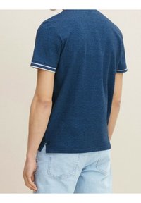 Tummansininen lyhythihainen paita, joka on valmistettu pehmeästä kankaasta ja jossa on raitakoristeiset hihansuut sekä klassinen leikkaus, esitettynä takaa.