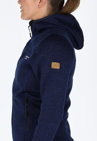 Giacca felpata con cappuccio di colore blu navy, presenta un motivo testurizzato, tasca laterale con zip e una toppa con logo marrone sulla manica. Include un colletto alto.