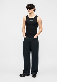 Jeune homme portant un débardeur noir avec une inscription blanche, un jean large foncé, des chaussures en cuir noires et des lunettes de soleil noires oversize, les mains dans les poches.