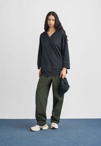 Μαύρο μακρυμάνικο τοπ με διαγώνιο ντεκολτέ και ριμπ υφή, συνδυασμένο με ελιάς πράσινο παντελόνι φαρδιάς γραμμής και λευκά αθλητικά παπούτσια. Κρατάει μαύρο καπέλο.