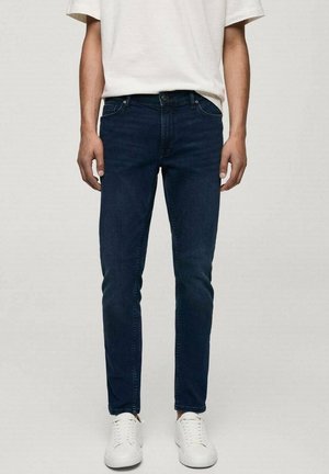 Uomo che indossa jeans aderenti blu scuro, sneakers bianche e una camicia bianca a maniche corte, in piedi davanti a uno sfondo neutro.