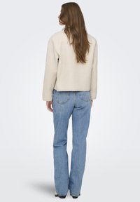 Kvinne med langt brunt hår står med ryggen til, iført en kremfarget, strukturert jakke og lyseblå, rette jeans med svarte sko.