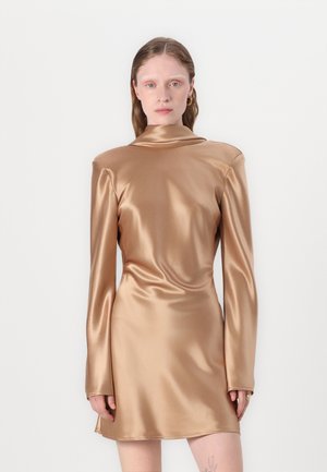 Kobieta o prostych brązowych włosach, ubrana w błyszczącą, brązową, mini sukienkę z długim rękawem i poduszkami na ramionach, stoi na tle jasnego, jednolitego tła.