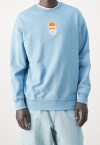 Sudadera azul claro hecha de tela suave, con un cuello redondo, puños acanalados y un logo bordado central en naranja y blanco.