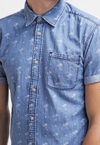 Camicia in denim a maniche corte di colore azzurro chiaro con motivo di palme e pois bianchi. Presenta una tasca sul petto e bottoni in plastica.