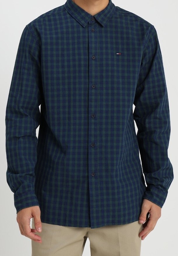Homme portant une chemise à carreaux bleu marine et verte à manches longues, boutonnée, et un pantalon beige, debout avec les mains détendues.