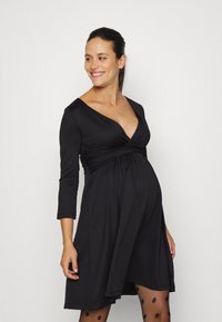Vestido de maternidade preto com mangas três-quartos, decote drapeado e cintura império. Meias-calças com padrão complementam o conjunto.