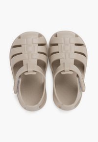Sandalias de plástico beige con la puntera cerrada, los lados abiertos y una correa ajustable en el talón. La suela presenta un patrón texturizado en espiral.
