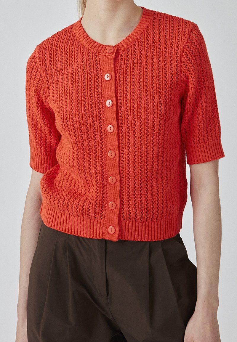 Cardigan en maille orange à manches courtes avec fermeture à boutons, porté avec un pantalon plissé taille haute de couleur marron foncé sur une personne debout.