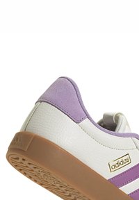 Zapatilla blanca con acentos de ante lavanda, cuero texturizado y suela de goma. Presenta un logo dorado y tres rayas en el lateral.