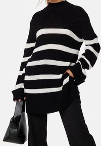 Schwarz-weiß gestreifter Strickpullover mit hohem Ausschnitt, langen Ärmeln und seitlichen Taschen. Model hält eine schwarze Handtasche.