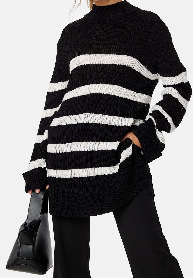 Schwarz-weiß gestreifter Strickpullover mit hohem Ausschnitt, langen Ärmeln und seitlichen Taschen. Model hält eine schwarze Handtasche.