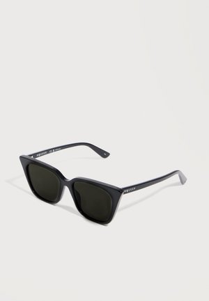 Lunettes de soleil noires angulaires avec des montures épaisses et des verres foncés, reposant sur une surface claire affichant la marque "McQueen" sur les branches.