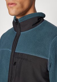 Chaqueta de forro polar con parte superior en verde azulado y sección inferior en negro, presenta un cierre de cremallera y un logo bordado. Textura suave y diseño de cuello alto.