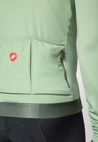 Light green cycling jacket features a zippered pocket, lightweight fabric, and a dark green hem. Red logo on the left side adds contrast.