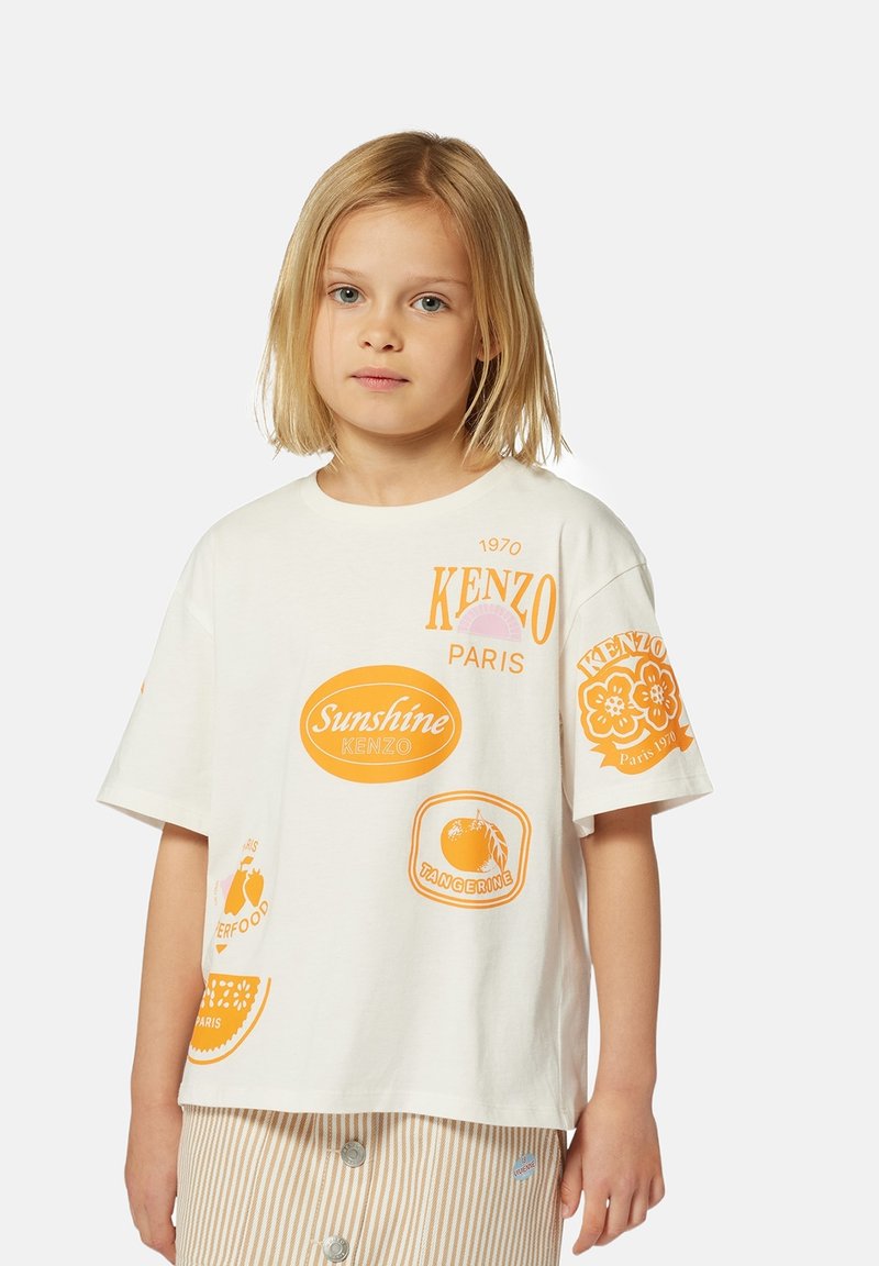 T-shirt en coton blanc avec des design imprimés orange, comprenant du texte et des logos, à manches courtes et à coupe décontractée.