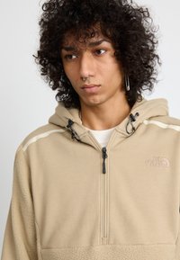Chaqueta de forro polar beige con capucha, cierre de cremallera y detalles en negro. Presenta una sección inferior texturizada y un logo bordado en el pecho.