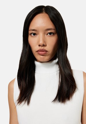 Jonge vrouw met lang donker haar, gekleed in een witte mouwloze coltrui, kijkt recht vooruit tegen een effen lichte achtergrond.