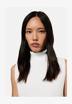 Joven con el pelo largo y oscuro, vistiendo un jersey blanco sin mangas de cuello vuelto, mirando de frente con un fondo claro y liso.