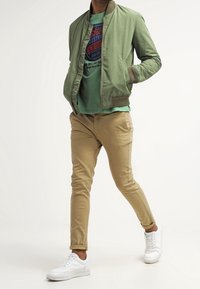 Zelená bomber bunda, světle modré tričko s grafickým potiskem, béžové slim-fit kalhoty a bílé tenisky. Neformální outfit s různými texturami.