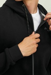 Felpa nera con zip caratterizzata da una texture liscia, cordini regolabili e un rivestimento interno bianco, parzialmente aperta per rivelare una t-shirt.