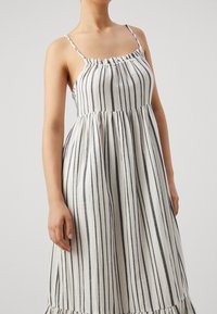 Robe blanche avec des rayures verticales noires, dotée de fines bretelles, d'un décolleté froncé et d'une taille évasée, finie par un ourlet à volants.