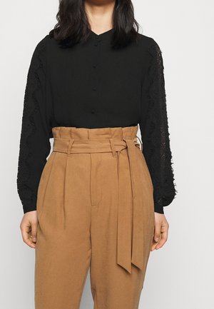 Czarna bluzka z koronkowymi zdobieniami na rękawach, w połączeniu z beżowymi, wysokim stanem spodniami z wiązanym paskiem i plisowanym przodem.