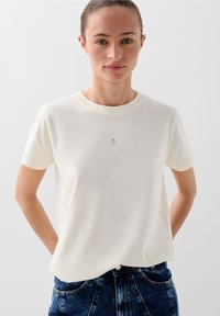 Cremefarbenes Baumwoll-T-Shirt mit kurzen Ärmeln und rundem Ausschnitt, ausgestattet mit einem kleinen gestickten Logo auf der Brust. Glatte Textur, lässige Passform.