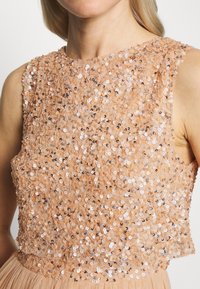 Beige säihkedes ülaosa kõrge kaelusega ja ilma varrukateta disainiga. Omab tekstuuriga pinda, millel on erineva suurusega ringikujulised sädelused.