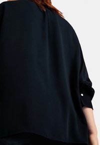 Schwarze, lockere Bluse aus glattem Material mit hochrollbaren Ärmeln und abgerundetem Saum. Sie verfügt über ein dezentes Knopfdetail an den Manschetten.