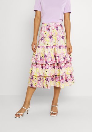 Mujer con falda floral de capas en tonos rosa, morado y amarillo, con una camiseta de manga corta lila clara y sandalias blancas con tacón.