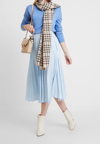 Ljust blå plisserad kjol i kombination med en blå tröja, rutigt halsduk och krämfärgade ankelboots. Accessoar: beige axelväska med strukturerad yta.