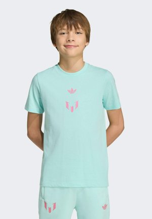 Niño pequeño con cabello castaño claro, vistiendo una camiseta Adidas azul claro y pantalones cortos a juego con logos rosas, sonriendo con las manos detrás de la espalda.