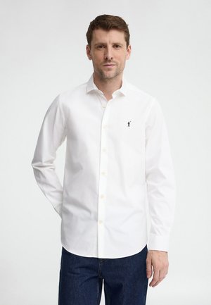 Mann trägt ein weißes langärmliges Hemd mit Knöpfen und einem kleinen eingestickten Logo, kombiniert mit dunkelblauen Jeans, stehend vor einem schlichten Hintergrund.