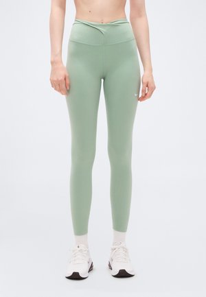 Žena v svetlozelenej legínach s vysokým pásom a bielym logom Nike, v kombinácii s bielymi ponožkami a bielymi športovými topánkami, stojaca pred bielym pozadím.