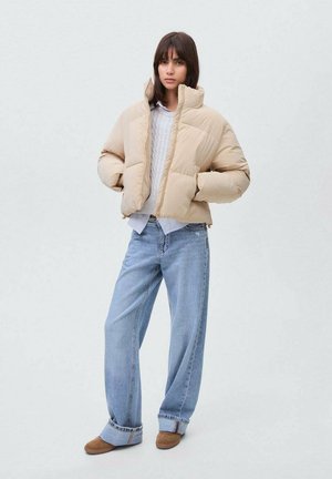 Frau trägt eine beigefarbene Steppjacke, einen weißen Pullover, weite hellblaue Jeans und braune Schuhe. Sie steht mit den Händen in den Taschen vor einem schlichten Hintergrund.
