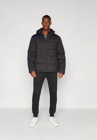 Άντρας στέκεται πρόσωπο με πρόσωπο φορώντας μια μαύρη γεμιστή ζακέτα με κουκούλα, μαύρο παντελόνι και λευκά αθλητικά παπούτσια μπροστά σε λευκό φόντο.