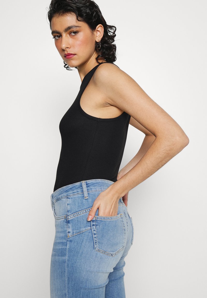 Junge Person mit dunklem lockigem Haar, die ein schwarzes ärmelloses Oberteil und hellblaue Jeans trägt, mit einer Hand in der Gesäßtasche, seitlich stehend.