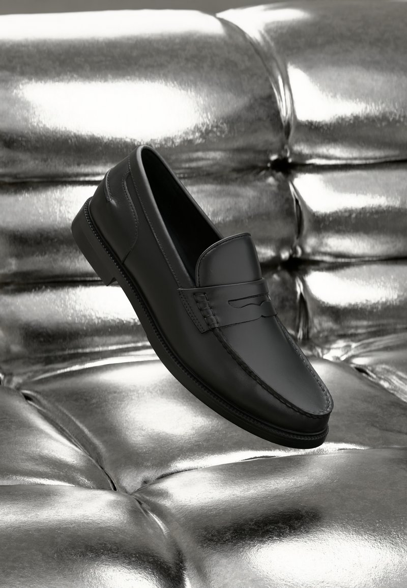 Mocassins en cuir noir à texture lisse, avec des détails de couture et un design classique, présentés sur un fond métallique et brillant.