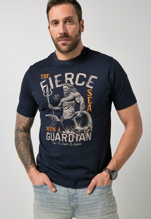 Mężczyzna z tatuażem na ramieniu, ubrany w granatową koszulkę z nadrukiem muskularnego syrena trzymającego trójząb, z napisem „Dziki morze potrzebuje opiekuna.”